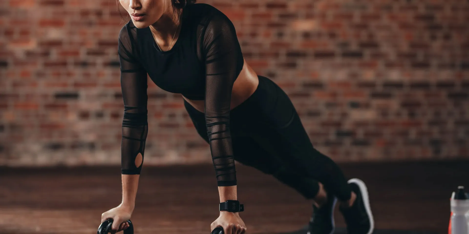 Sportliche Frau führt Liegestütze auf zwei Kettlebells im Plank-Stütz vor einer Backsteinwand aus