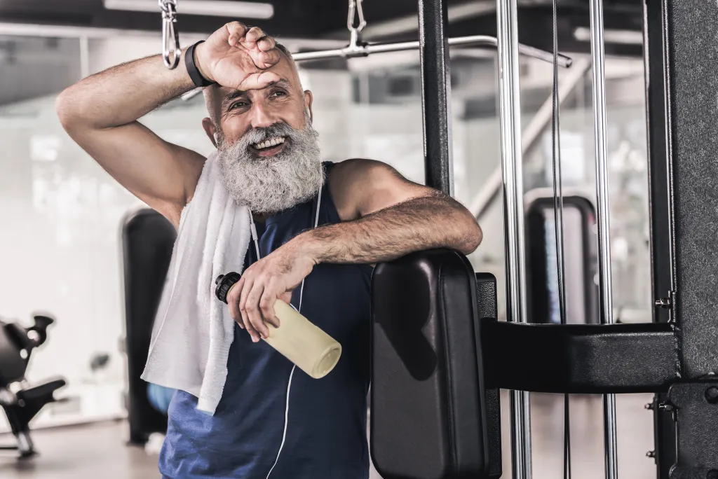 Lächelnder älterer Mann mit grauem Vollbart wischt sich nach dem Training im Fitnessstudio den Schweiß von der Stirn