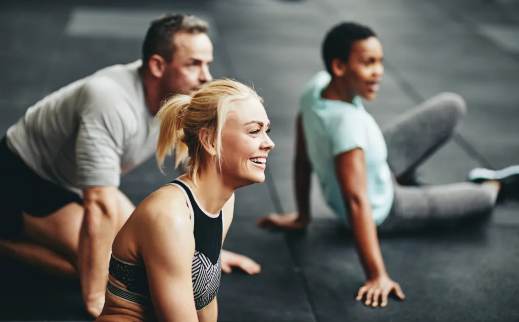 Lächelnde blonde Frau sitzt mit ihrer Sportgruppe während einer Trainingspause auf dem Boden des Fitnessstudios